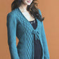 A woman with long, dark hair is wearing a stylish teal cardigan with decorative patterns from the "No-Sew Knits" collection by Ingram Content. The cardigan is tied at the front over her black dress. She stands against a plain, light brown background and has a slight smile on her face—a perfect example of trendy styling.