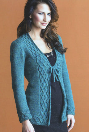 A woman with long, dark hair is wearing a stylish teal cardigan with decorative patterns from the "No-Sew Knits" collection by Ingram Content. The cardigan is tied at the front over her black dress. She stands against a plain, light brown background and has a slight smile on her face—a perfect example of trendy styling.