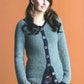 A woman with long, wavy hair stands against an orange background, wearing the No-Sew Knits gray cardigan by Ingram Content, featuring wooden buttons and dark trim. She has her hands in the pockets of her blue jeans and is smiling slightly. Her trendy styling is complemented by dangling earrings and a comfortable sweater.