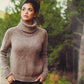 A young woman with dark hair tied up, wearing a cozy brown turtleneck sweater from Plum Dandi Knits by Ingram Content and black pants, stands outdoors with her hands in her pockets. She's looking to the side against a backdrop of lush greenery and trees.