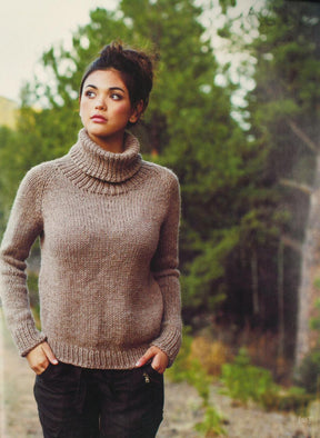 A young woman with dark hair tied up, wearing a cozy brown turtleneck sweater from Plum Dandi Knits by Ingram Content and black pants, stands outdoors with her hands in her pockets. She's looking to the side against a backdrop of lush greenery and trees.