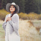 A woman stands outdoors near a grassy and wooded area, wearing a wide-brimmed hat and a cozy thick wool shawl from Plum Dandi Knits over a grey top. She looks directly at the camera with a calm expression, and the background features blurred autumn foliage and a small body of water. This beautiful shawl is crafted by Ingram Content.