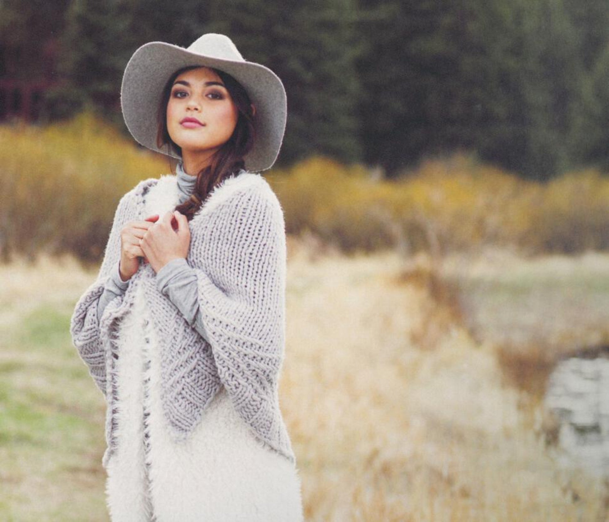 A woman stands outdoors near a grassy and wooded area, wearing a wide-brimmed hat and a cozy thick wool shawl from Plum Dandi Knits over a grey top. She looks directly at the camera with a calm expression, and the background features blurred autumn foliage and a small body of water. This beautiful shawl is crafted by Ingram Content.