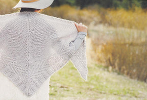 A person with long hair, donning a wide-brimmed hat and a long-sleeved shirt, showcases a luxurious knit shawl from the Plum Dandi Knits collection by Ingram Content in an outdoor setting. The scenery features dry vegetation and softly focused grassy, sunlit fields.