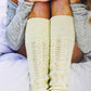 A person with long hair and a soft smile sits with their knees drawn up and hands resting on their knees, wearing a light gray long-sleeve top and cream-colored knitted leg warmers from Plum Dandi Knits by Ingram Content. The luxurious knits add to the cozy, relaxed outdoor atmosphere in the background.