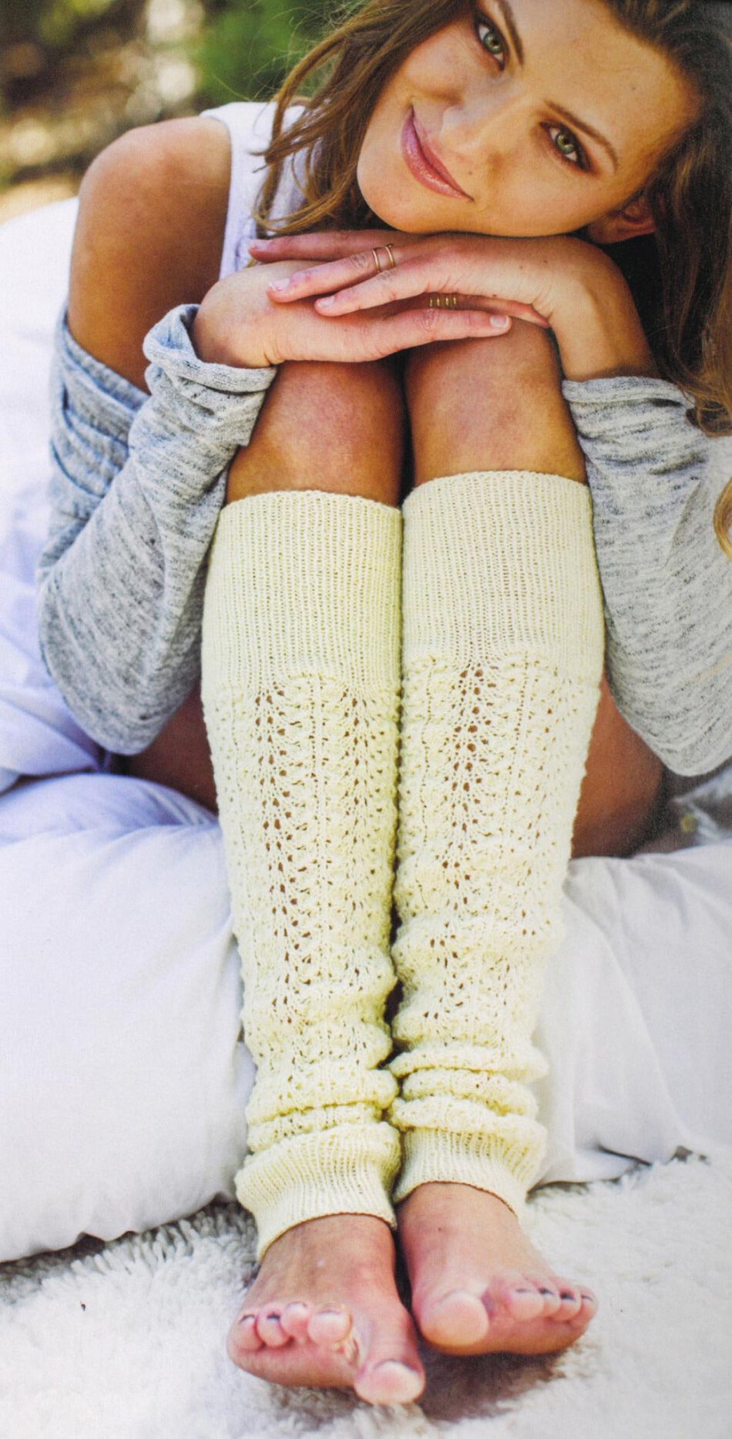 A person with long hair and a soft smile sits with their knees drawn up and hands resting on their knees, wearing a light gray long-sleeve top and cream-colored knitted leg warmers from Plum Dandi Knits by Ingram Content. The luxurious knits add to the cozy, relaxed outdoor atmosphere in the background.