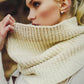 A person with light blonde hair is partially covering their face with a luxurious, chunky off-white knit scarf from Plum Dandi Knits by Ingram Content. They are wearing a beige coat and have a gold triangular earring visible. The background is blurred, suggesting an outdoor setting.