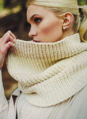 A person with light blonde hair is partially covering their face with a luxurious, chunky off-white knit scarf from Plum Dandi Knits by Ingram Content. They are wearing a beige coat and have a gold triangular earring visible. The background is blurred, suggesting an outdoor setting.