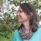 A woman with shoulder-length brown hair, wearing a turquoise cardigan and a multicolored scarf made from Noema yarn, smiles at a blooming tree adorned with pink flowers. The backdrop is a serene outdoor setting with lush greenery and vibrant blossoms, offering the perfect inspiration for projects found in the Noema Book by Louisa Harding from Knitting Fever / Euro Yarns.