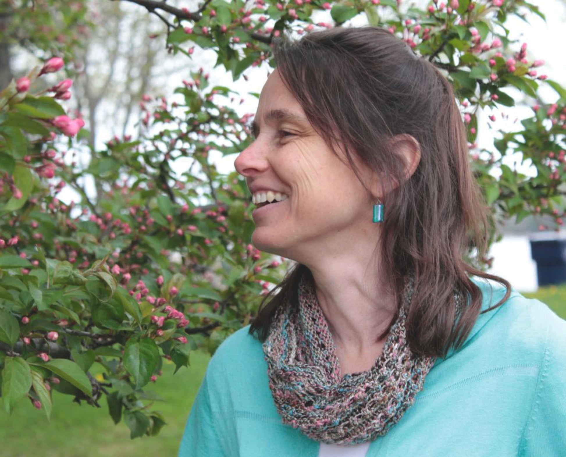 A woman with shoulder-length brown hair, wearing a turquoise cardigan and a multicolored scarf made from Noema yarn, smiles at a blooming tree adorned with pink flowers. The backdrop is a serene outdoor setting with lush greenery and vibrant blossoms, offering the perfect inspiration for projects found in the Noema Book by Louisa Harding from Knitting Fever / Euro Yarns.