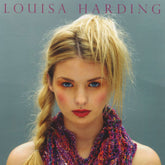 A woman with blonde hair styled in a high ponytail and braid, wearing vibrant makeup including bold eyeshadow and red lipstick, gazes directly at the camera. She is dressed in a colorful, textured knit garment made from Noema yarn. Above her head, the text "LOUISA HARDING" celebrates knitting projects from the Noema Book by Knitting Fever / Euro Yarns.