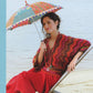 A woman sits on a deck chair by the water, holding a colorful, patterned umbrella. She is wearing a red and pink chevron-patterned top crafted from Knitting Fever / Euro Yarns' Noro Favourites Silk Garden Sock Yarn, paired with a red skirt and a long necklace featuring a peace sign pendant. Her hair is styled up, and she has a relaxed, content expression.