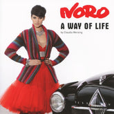 A model with short dark hair poses in a vibrant red dress paired with a striped cardigan crafted from Noro yarns. She stands beside a vintage black car, exuding magical color and elegance. The text reads "Noro A Way of Life" by Knitting Fever / Euro Yarns and Claudia Wersing.