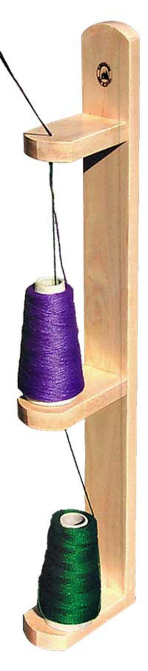 The Leclerc Doubling Stand by Leclerc Looms is a vertical wooden thread rack mounted on the wall, holding two large spools of thread. The top spool is purple and the bottom spool is green. Each spool feeds thread upwards through individual slots in the rack, making it perfect for weaving or twisting yarns effortlessly.