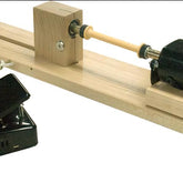 The Leclerc Double-Ended Electric Bobbin Winder by Leclerc Looms features a black motor on the right end mounted on a wooden base. A foot pedal connected to the machine is positioned to the left, used for controlling its operation. The cylindrical wooden bobbin is set up between two centers on the winder.