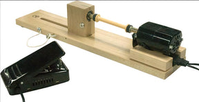 The Leclerc Double-Ended Electric Bobbin Winder by Leclerc Looms features a black motor on the right end mounted on a wooden base. A foot pedal connected to the machine is positioned to the left, used for controlling its operation. The cylindrical wooden bobbin is set up between two centers on the winder.