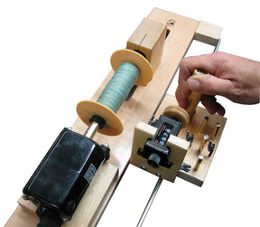 A close-up of the Leclerc Double-Ended Electric Bobbin Winder by Leclerc Looms, showcasing spools of green thread. A person adjusts a knob with their right hand. The device features a counter display and various visible mechanical components, including a black motor and metal rods.