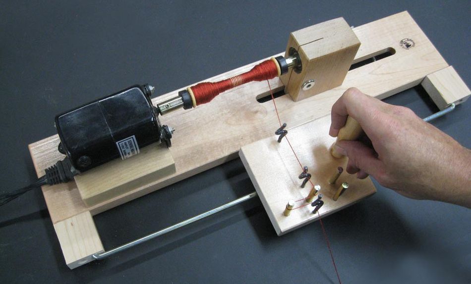 A hand operates a Leclerc Double-Ended Electric Bobbin Winder by Leclerc Looms. The machine has a motor on one end and a spool of red wire on the other, with several pegs guiding the wire as it is wound onto the spool.