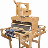 The Leclerc Diana 24" 16-Shaft, Computer Dobby Floor Loom by Leclerc Looms is a traditional wooden loom used for weaving textiles. It features a sturdy frame with various levers and mechanisms, and a partially woven fabric with blue and white stripes is visible on the loom.