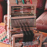 A Leclerc Voyageur Table Loom by Leclerc Looms in action, with black threads being woven through it. Several colorful levers are visible on top. The portable loom is positioned on a colorful woven mat, with some blurred background furniture.