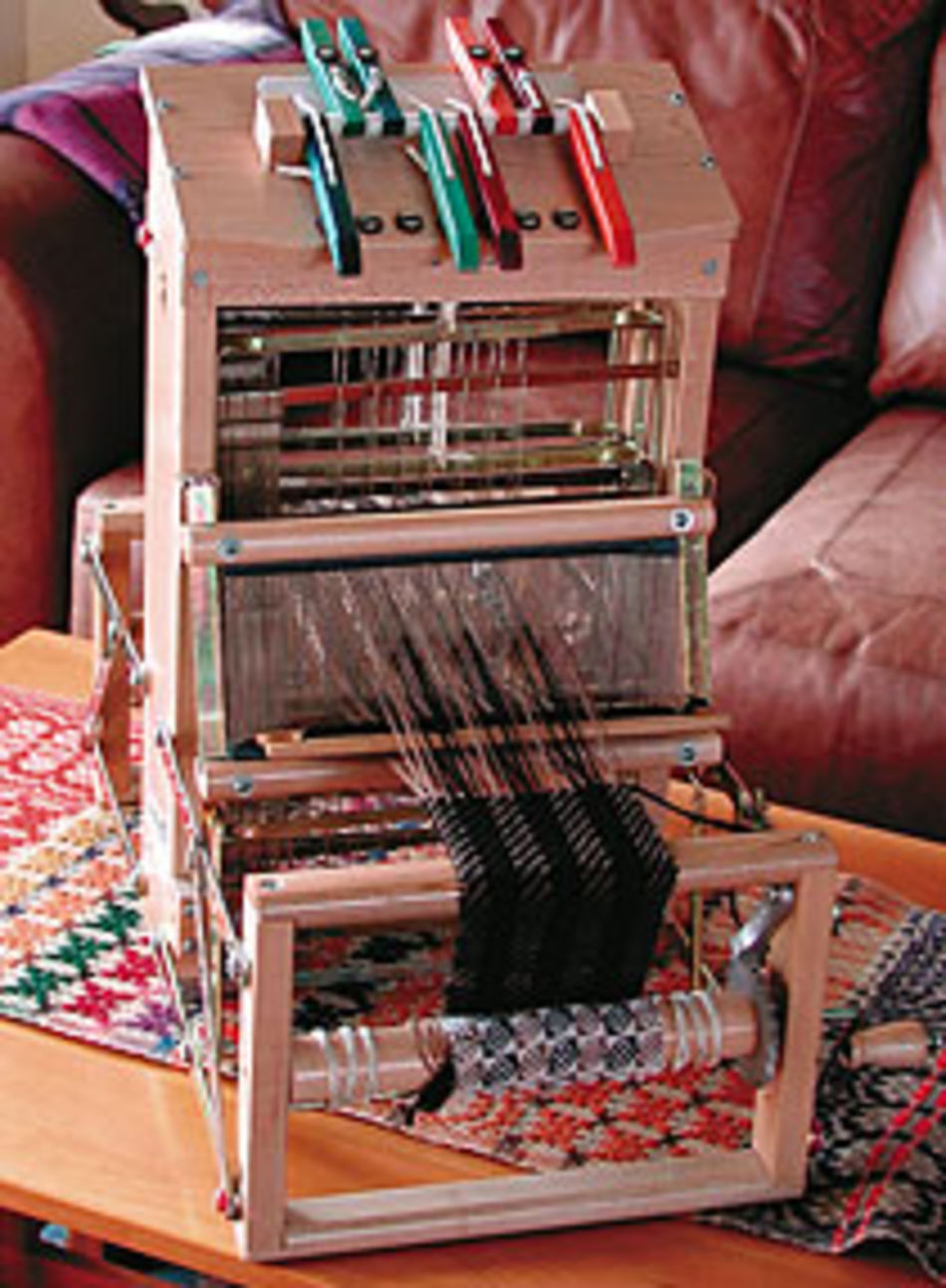 A small, intricate Leclerc Voyageur Table Loom from Leclerc Looms is set up on a wooden table, adorned with colorful clothespins on top. Various threads are strung through the machine, while a woven, patterned fabric emerges at the bottom. In the background, a brown couch with a textured blanket draped over it can be seen.