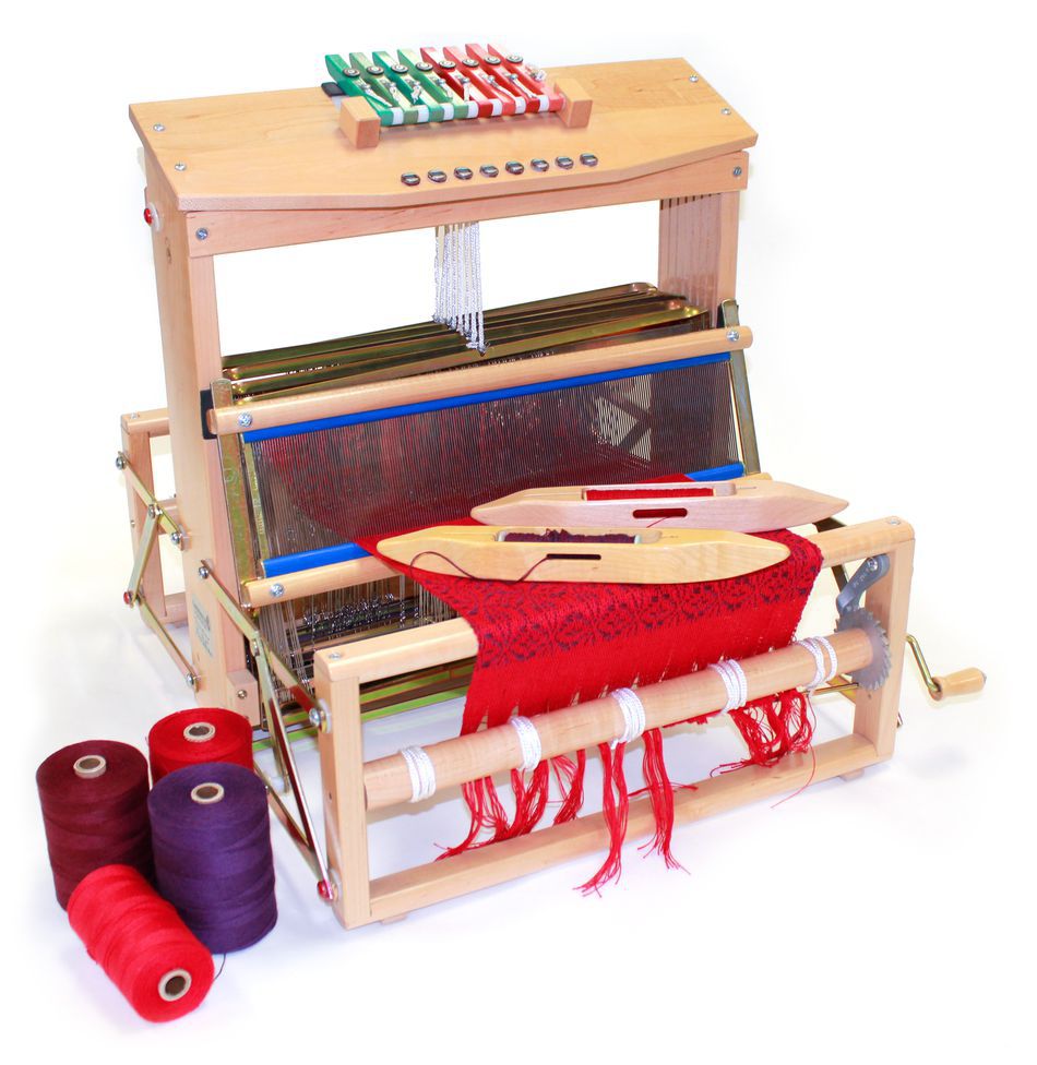 A Leclerc Voyageur Table Loom by Leclerc Looms, complete with a partially woven red textile and two weaving shuttles in the middle. Rolls of thread in red, purple, and brown are positioned beside the loom. This portable loom features multiple levers and threads in various colors, making it perfect for creative projects on the go.