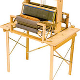 The Leclerc Table Loom Stand for Dorothy or Voyageur is a versatile wooden tabletop loom available in 4 or 8 shaft variants, ideal for handcrafting textiles and fabrics. It features multiple colored threads and intricate weaving mechanisms, supported by a sturdy wooden frame with aligned strings and gears. Additionally, the loom includes convenient side shelves for tool storage but does not come with treadles.