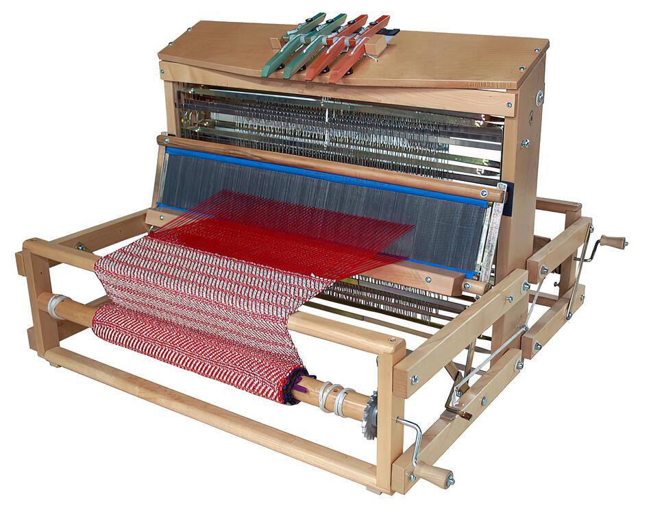 A Leclerc Voyageur Table Loom by Leclerc Looms with a red and white striped fabric in progress. The loom features several heddles, beams, and levers, along with colored shuttles on top. The intricate mechanisms and threads are visible, showcasing the traditional weaving process.