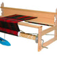 The Leclerc Bergere Rigid Heddle Loom, 24" by Leclerc Looms showcases a partially woven black and red fabric. The wooden frame supports various rods and levers, with ratchet brakes ensuring precision. A blue shuttle rests on the loom, while metal components adjust tension and fabric positioning.