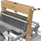 A close-up view of a Leclerc Dorothy 4-shaft section by Leclerc Looms in operation, showing various threads being woven into fabric. The wooden frame holds metal parts and side crossmembers, with colored handles likely for adjusting the machine. The loom is partially assembled with intricate detailing.