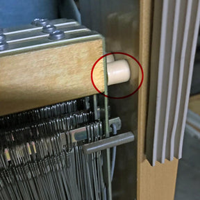 A close-up of a Leclerc loom with metal heddles and a Leclerc Shaft Frame End (beige plastic) circled in red on the side. Various tensioning devices and end pieces are visible. The loom, ideal for replacing lost or damaged components, is partially obscured by nearby objects.
