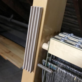 A close-up of part of a wooden loom, highlighting the heddles and a vertical comb-like structure attached to the frame. The slightly blurred background hints at more sections of the Leclerc Floor Loom and some wooden beams, with Leclerc Rubber Beater Bumper Pads for Floor Looms by Leclerc Looms discreetly in place.