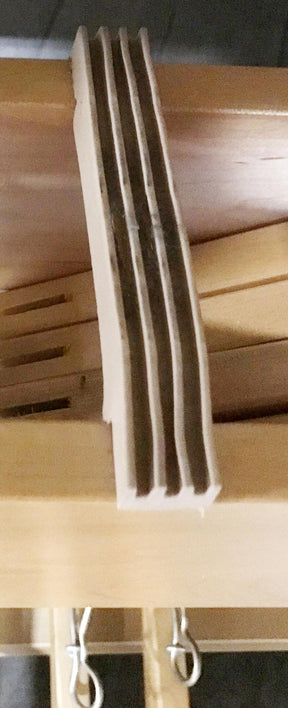 A close-up image of a wooden slat made from several thin layers of wood glued together. The multi-layered, laminated structure is placed on a wooden surface. In the background, Leclerc Rubber Bumper Under Shaft Pads for Floor Looms by Leclerc Looms are visible, helping to absorb impact and reduce noise.