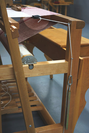 A close-up view showcases a Leclerc Clip Temple by Leclerc Looms, with a thread running vertically through it. The loom is partially covered with fabric, while the foreground reveals weaving accessories such as a metal rod attached to the thread and the wooden frame. In the background, more wooden structures are visible.