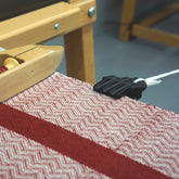 A close-up of a Leclerc loom in action. The fabric being woven has a classic herringbone pattern with a bold red stripe running down the middle. A shuttle with thread is visible, and a Leclerc Clip Temple from Leclerc Looms is attached to the white cord of the loom.