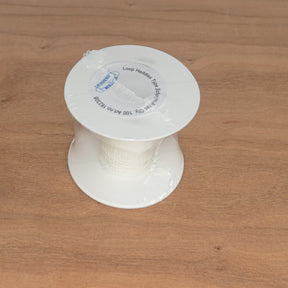 A spool of white Texsolv Heddles from Leclerc Looms, designed for the Schacht Arras Tapestry Loom with a 4" specification and resembling a continuous chain, is wrapped in clear plastic and rests on a wooden surface.