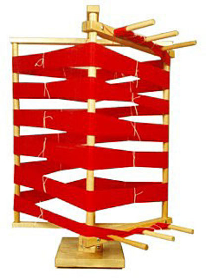 A Leclerc Warping Reel - vertical by Leclerc Looms stands laminated with a red yarn, winding in a zigzag pattern between the wooden supports. The warping reel is positioned upright on a square base with multiple pegs extending horizontally to hold the yarn.