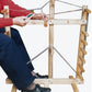 A person wearing a red sweater and black shoes is using the Leclerc Cendrel Standing Inkle Loom/Warping Frame from Leclerc Looms, holding a tool in their right hand. The frame includes several dowels and metal rods, which are used for weaving and repairing chair seats.