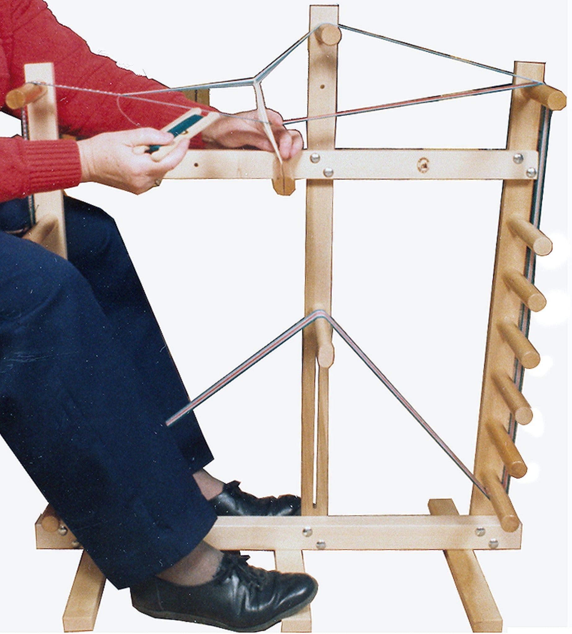 A person wearing a red sweater and black shoes is using the Leclerc Cendrel Standing Inkle Loom/Warping Frame from Leclerc Looms, holding a tool in their right hand. The frame includes several dowels and metal rods, which are used for weaving and repairing chair seats.