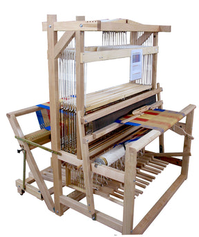 The Leclerc Colonial V2 Jack Floor Loom by Leclerc Looms is a wooden handloom featuring multiple shafts and heddles, displaying woven fabric in progress. The structure includes pedals at the bottom and a partially completed patterned textile with multicolored threads, showcasing the intricate weaving process.