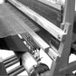 A black and white image of the Leclerc Batten Weighted Beater in action. The loom, crafted by Leclerc Looms, features wooden frames with a partly woven fabric emerging from it. The warp threads are tightly stretched, and the weft threads are visible on the partially finished cloth.