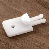 The Texsolv 2-Legged Anchor Pegs by Glimakra are small white plastic components featuring a rectangular base, a circular hole near one end, and extended double prongs at the other. One prong has a larger rounded tip. These components are pictured on a wooden surface and resemble elements often found in Texsolv systems or used for harness tie-ups.