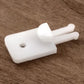 The Texsolv 2-Legged Anchor Pegs by Glimakra are small white plastic components featuring a rectangular base, a circular hole near one end, and extended double prongs at the other. One prong has a larger rounded tip. These components are pictured on a wooden surface and resemble elements often found in Texsolv systems or used for harness tie-ups.