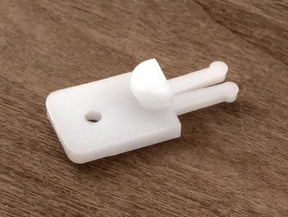 The Texsolv 2-Legged Anchor Pegs by Glimakra are small white plastic components featuring a rectangular base, a circular hole near one end, and extended double prongs at the other. One prong has a larger rounded tip. These components are pictured on a wooden surface and resemble elements often found in Texsolv systems or used for harness tie-ups.