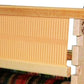 A close-up of the Leclerc 24" Bergere Rigid heddle Reed by Leclerc Looms, crafted from light-colored wood and metal. The reed, featuring evenly spaced vertical slots, occupies most of the frame. The lower part of the loom with warp threads is partially visible.