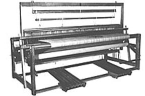 The black and white image showcases a Leclerc Kebec II 4-Shaft Floor Loom from Leclerc Looms. This old-fashioned weaving device features a complex assembly of frames, beams, rods, and a large fabric roll centrally positioned. It is designed for weaving textiles by interlacing threads either manually or mechanically.