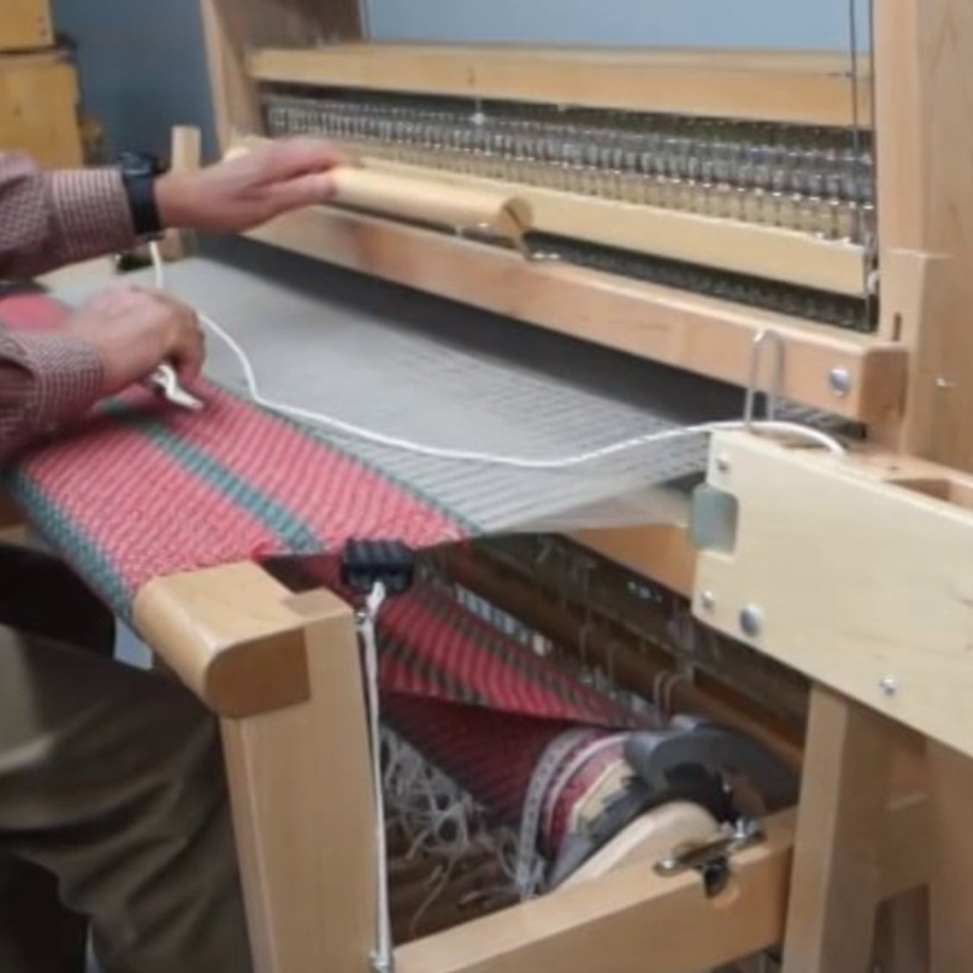 A person is using a Leclerc Flying Shuttle Beater by Leclerc Looms to weave a red and green patterned textile in one of the cozy weaving studios. Seated with one hand on the loom's bar, strings and threads are clearly visible. The Leclerc Flying Shuttle Beater, equipped with various mechanical parts, including a flying shuttle, facilitates the intricate work.