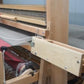 A person using the Leclerc Flying Shuttle Beater from Leclerc Looms in a weaving studio to create wide fabrics with red, green, and white threads. One hand holds the tool pushing horizontal threads into place, while the other maneuvers the fabric. The loom’s heddles and reeds are visible, showcasing the intricate weaving process.