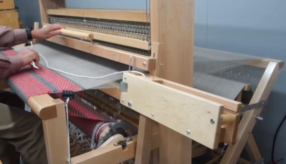 A person using the Leclerc Flying Shuttle Beater from Leclerc Looms in a weaving studio to create wide fabrics with red, green, and white threads. One hand holds the tool pushing horizontal threads into place, while the other maneuvers the fabric. The loom’s heddles and reeds are visible, showcasing the intricate weaving process.