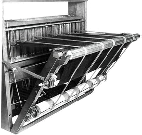 A monochrome image of an early mechanical loom, specifically the Leclerc Double Warp Beam Standard by Leclerc Looms. The loom features an array of warp threads stretched vertically along with multiple rollers and beams. Various mechanical parts and gears are visible, showcasing the complex machinery used in weaving fabrics.